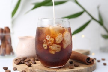 Pouring milk into tasty iced coffee in glass on white table, closeup