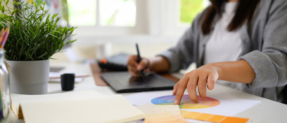 Cropped shot woman graphic designer creating logo on digital tablet in bright modern workspace with computer and color palette
