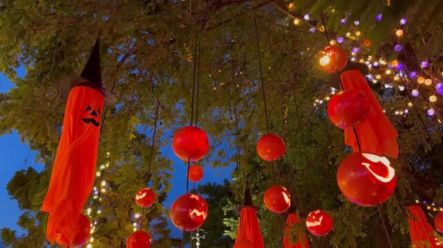 Orange cloaked ghosts and pumpkin devils hang from trees and spin in the wind, Halloween garden decorations.