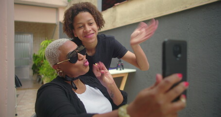 Smiling Black woman and teenage son taking a selfie together outdoors, waving at the camera, bonding in a joyful and warm family moment with greenery in the background