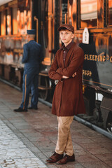 A thoughtful young man in a brown coat and a retro-style cap holds a rolled-up newspaper on a train...