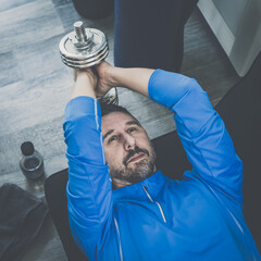 Determined sporty young man working out and trains at home. Fit male doing exercise with weights for biceps. Strong boy lifting the dumbbells in the living room. Wellness, health, sport, gym concept