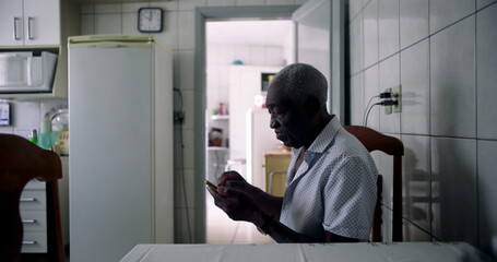 Elderly African American man seated at dining table in cozy kitchen, focused on using a smartphone, showcasing everyday moments of technology use in a homely atmosphere