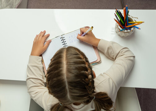 Overhead shot of young girl sitting at table and writing in notebook at home. Cozy educational setting ideal for themes of childhood, homeschooling, creativity, concentration. - Powered by Adobe