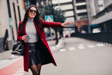 Chic woman in red coat and skirt hailing taxi outdoors in urban city street with smile and...