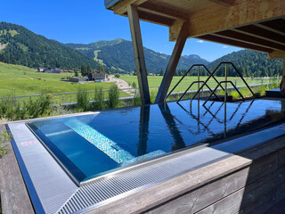 Infinity-Pool mit Bergblick in den Alpen &ndash; moderner Spa-Bereich mit Panoramablick auf gr&uuml;ne Wiesen, W&auml;lder und Berge unter blauem Himmel