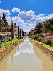 Historische Festungsmauer mit Wassergraben und Zypressen unter blauem Himmel in Palma de Mallorca, Spanien. Malerische Aussicht mit Wolken und Spiegelung im Wasser.
