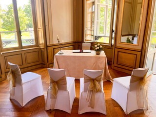 Elegant dekorierter Raum f&uuml;r eine standesamtliche Trauung mit vier wei&szlig;en St&uuml;hlen und goldenen Schleifen, festlich gedecktem Tisch, Kerzen und Blumen, in historischem Ambiente mit Holzvert&auml;felung.