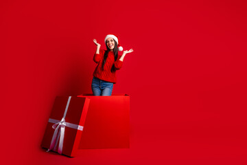 Young woman in festive Santa hat emerging from a giant gift box against red background, embracing...