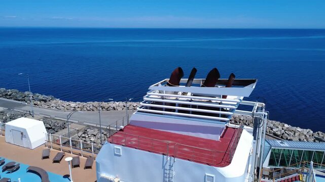 A view of the exhaust pipes of a cruise ship docked near a boarding bridge on the shore of a blue ocean that fades into the horizon. Matane, Quebec, Canada, 2025.