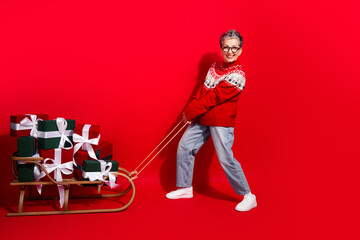 Festive woman pulling a gift-laden sleigh in cozy red sweater on bright red background, spreading holiday joy