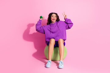 Young woman in vibrant sports attire sitting on exercise ball with cheerful expression on pink...