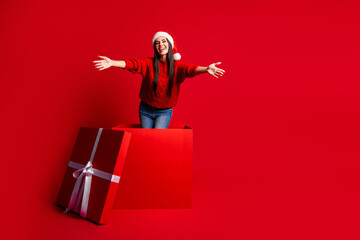 Festive woman in Santa hat emerging joyfully from large gift box on vibrant red background...