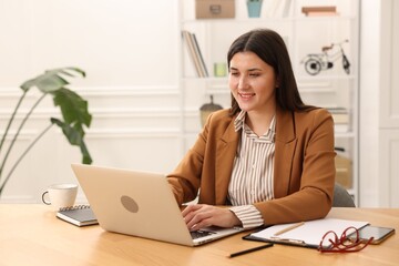 Smiling woman in suit working with laptop at wooden desk in home office. Space for text