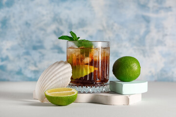 Glass of cold Cuba Libre cocktail with seashell and plaster podiums on white table against blue grunge wall