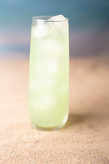 refreshing green cocktail in tall glass with ice on sandy surface against soft blue gradient background evoking beachside relaxation and tropical vibes, context, idyllic, appealing, exotic, elegant