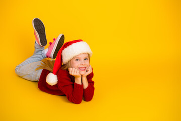 Playful Christmas kid in red sweater wearing a Santa hat lying on a bright yellow background dreaming of holiday joy and winter fun with youth and imagination