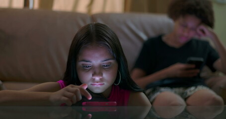 Young girl focused on smartphone lying on table with glowing light on face while teenager boy in...