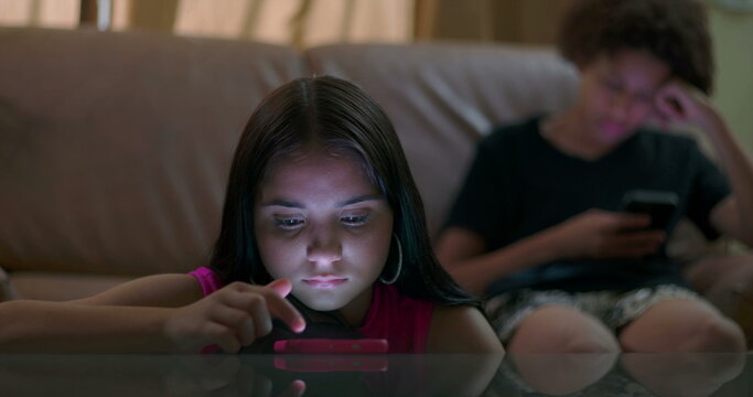 Young girl focused on smartphone lying on table with glowing light on face while teenager boy in background also on phone, portraying social media addiction and digital disconnection