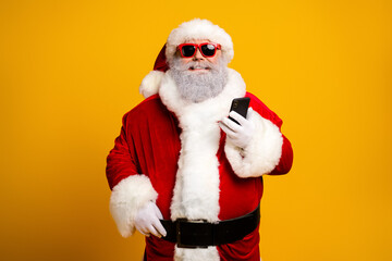 Cool Santa Claus in red costume with sunglass holding a smartphone against vibrant yellow backdrop, feeling modern