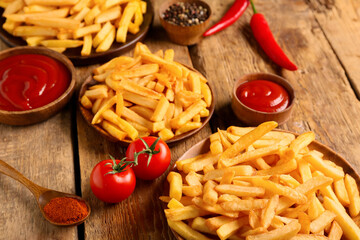 Plates with tasty french fries, bowls of ketchup, chili peppers, tomatoes and peppercorns on wooden background