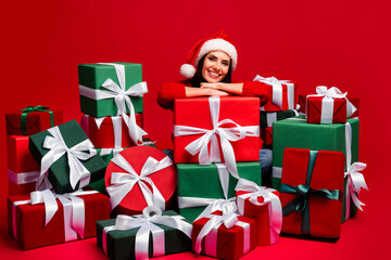 Cheerful young woman in a Santa hat surrounded by colorful gifts on a red background, celebrating the joy of Christmas