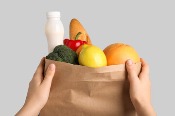 Female hands with vegetables, fruits, baguette and bottle of yogurt in paper bag on grey background