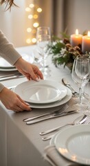 Woman arranging an elegant table setting with white plates, wine glasses, silverware, candles, and festive greenery decorations, creating a sophisticated holiday dining atmosphere.