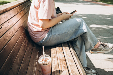 Person sitting on a wooden bench, enjoying a drink while checking their smartphone. Casual attire suggests a leisurely outdoor break, capturing a relaxed and tranquil mood.