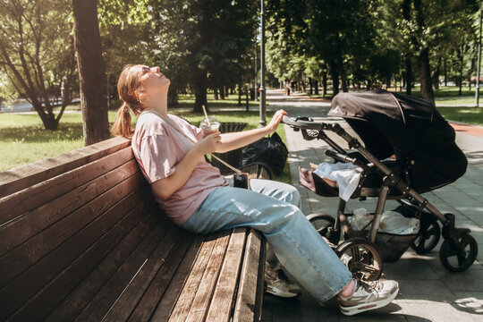 Woman enjoying a sunny day on a park bench, sipping a drink as a baby stroller is parked nearby. She leans back, soaking in the sun, taking a moment of self-care and relaxation.