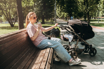 Woman enjoying a sunny day on a park bench, sipping a drink as a baby stroller is parked nearby. She leans back, soaking in the sun, taking a moment of self-care and relaxation.