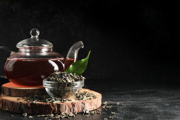 Composition with teapot of black tea and bowl of dry leaves on black grunge background, closeup