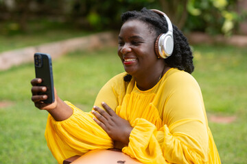 Expressive Black woman in a yellow dress and headphones waving during a video call on her smartphone outdoors