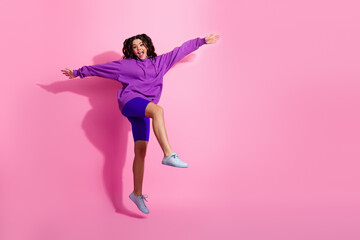 Vibrant young woman in stylish purple sportswear joyfully jumping against a pink background...
