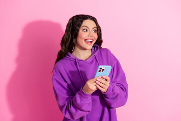 Cheerful young woman with phone in hand wearing purple hoodie on pink background
