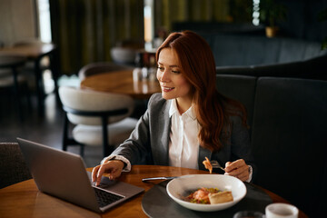 Happy businesswoman reading email on laptop while eating in restaurant.