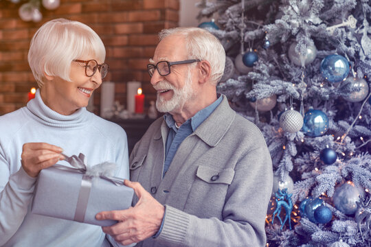 Senior man giving a christmas gift to his wife at home. Present gift sharing between elderly old couple spouses for New Year. Family time together, reunion for winter holiday - Powered by Adobe