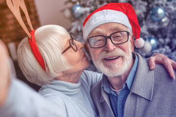 Happy smiling senior couple in hats taking selfie at home. Christmas celebration of elderly spouses grandparents vlogging blogging in front of fir tree