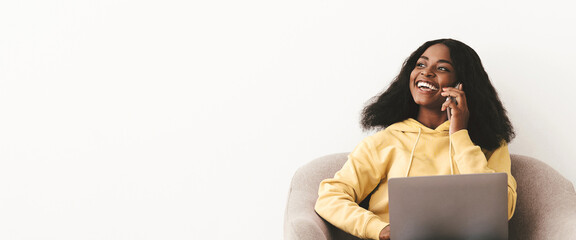 Happy pretty young black woman in casual sitting at arm chair by empty white wall, having phone...