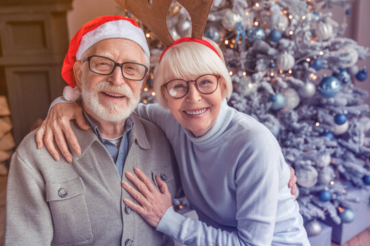 Portrait of happy smiling senior couple in santa hats celebrating at home. Elderly old grandparents hugging embracing cuddling for Christmas New Year Eve looking at camera together