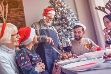 Grandfather in santa hat carving chicken at christmas dinner at home. New Year Eve celebration. Festive table with family reunion. Nutrition food for party, thanksgiving tradition