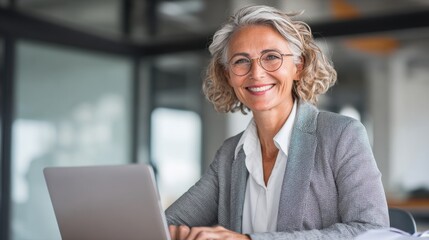 A confident businesswoman with gray hair is sitting at a desk in a bright office. She is smiling while typing on her laptop showcasing a positive work environment.