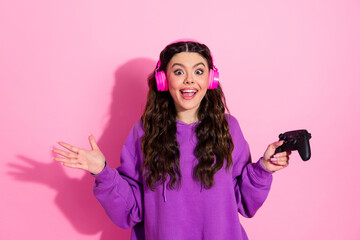 Energetic young woman in stylish pink headphones and purple sweatshirt posing with a gaming...