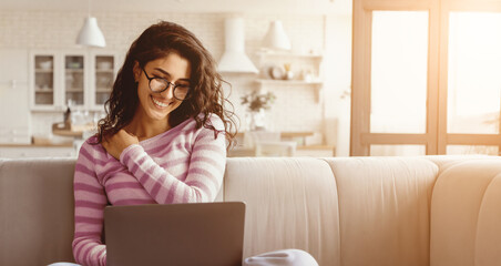 A young woman with curly hair sits on a comfortable sofa, happily interacting with her laptop. Sunlight streams through the window, creating a warm and inviting atmosphere.