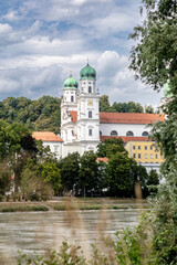 A summer view of St. Stephen&rsquo;s Cathedral in Passau, Germany, seen from the Innstadt side across the river Inn, surrounded by green trees and historic architecture under a cloudy sky.