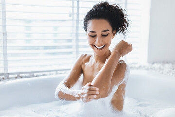 A woman is smiling as she relaxes in a bathtub filled with bubbles. Natural light fills the room, creating a peaceful atmosphere. She looks joyful and carefree.