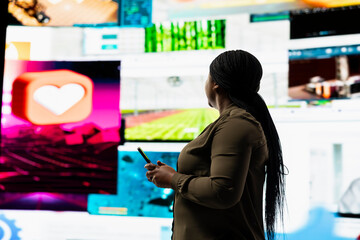 User engages with multimedia content on cellphone, sending likes across social networks. African american woman browses dashboard of streaming videos and social media feeds on digital wall