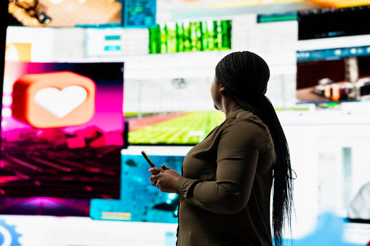 User engages with multimedia content on cellphone, sending likes across social networks. African american woman browses dashboard of streaming videos and social media feeds on digital wall - Powered by Adobe