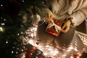 Happy woman holding stylish gift box at christmas tree with lights in evening close up,top view....