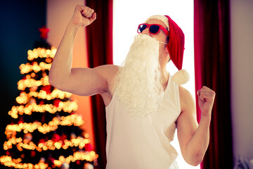 Santa Claus in a modern style celebrating fitness and Christmas, wearing sunglass and flexing near...